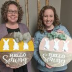 Two women holding wooden signs with bunnies on it and the words hello spring.