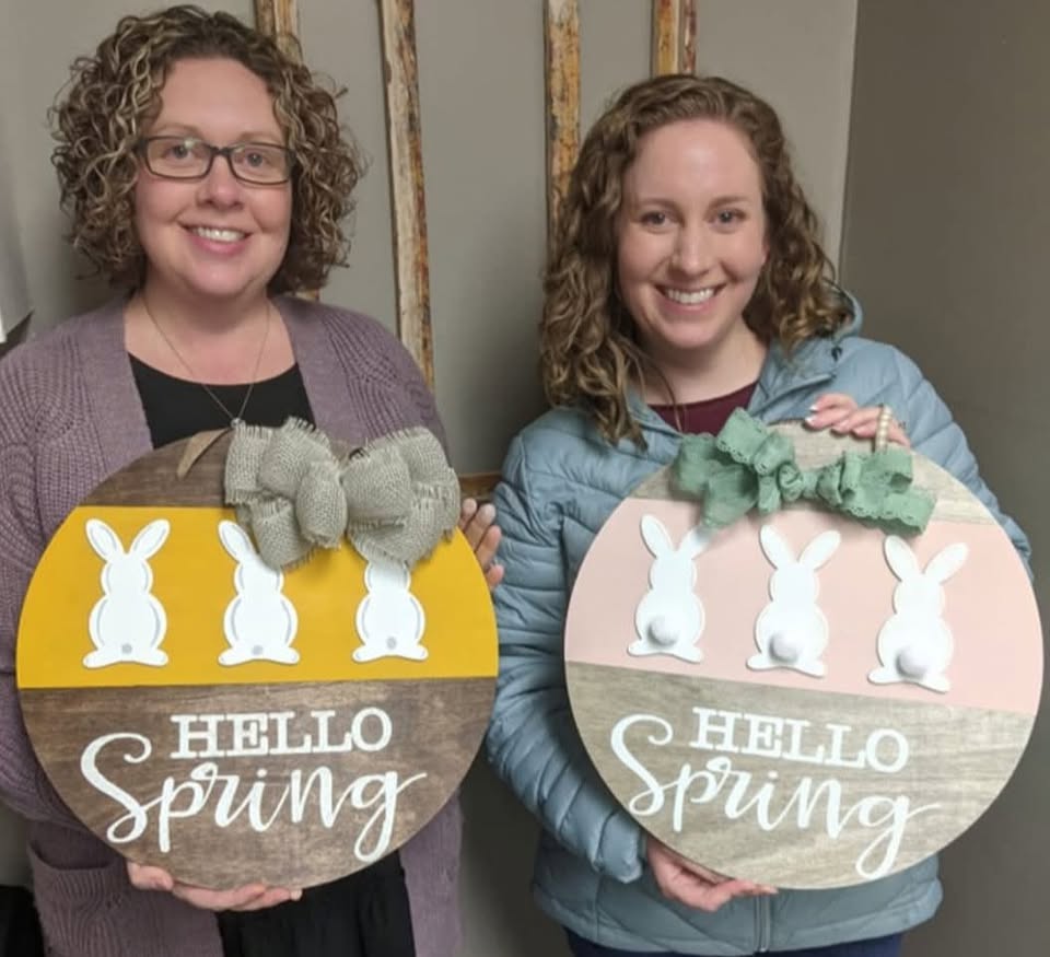 Two women holding wooden signs with bunnies on it and the words hello spring.