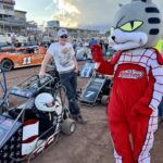 A small child in a race car is greeted by the Cedar lake Speedway mascot