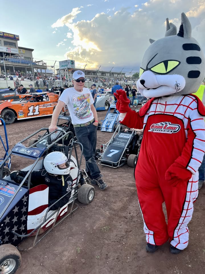 A small child in a race car is greeted by the Cedar lake Speedway mascot