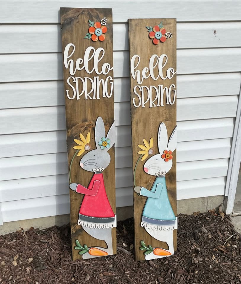 A photo of two bunny porch leaners the one of the left is a bunny wear a red dress, the one of the right is a bunny wearing a blue dress.