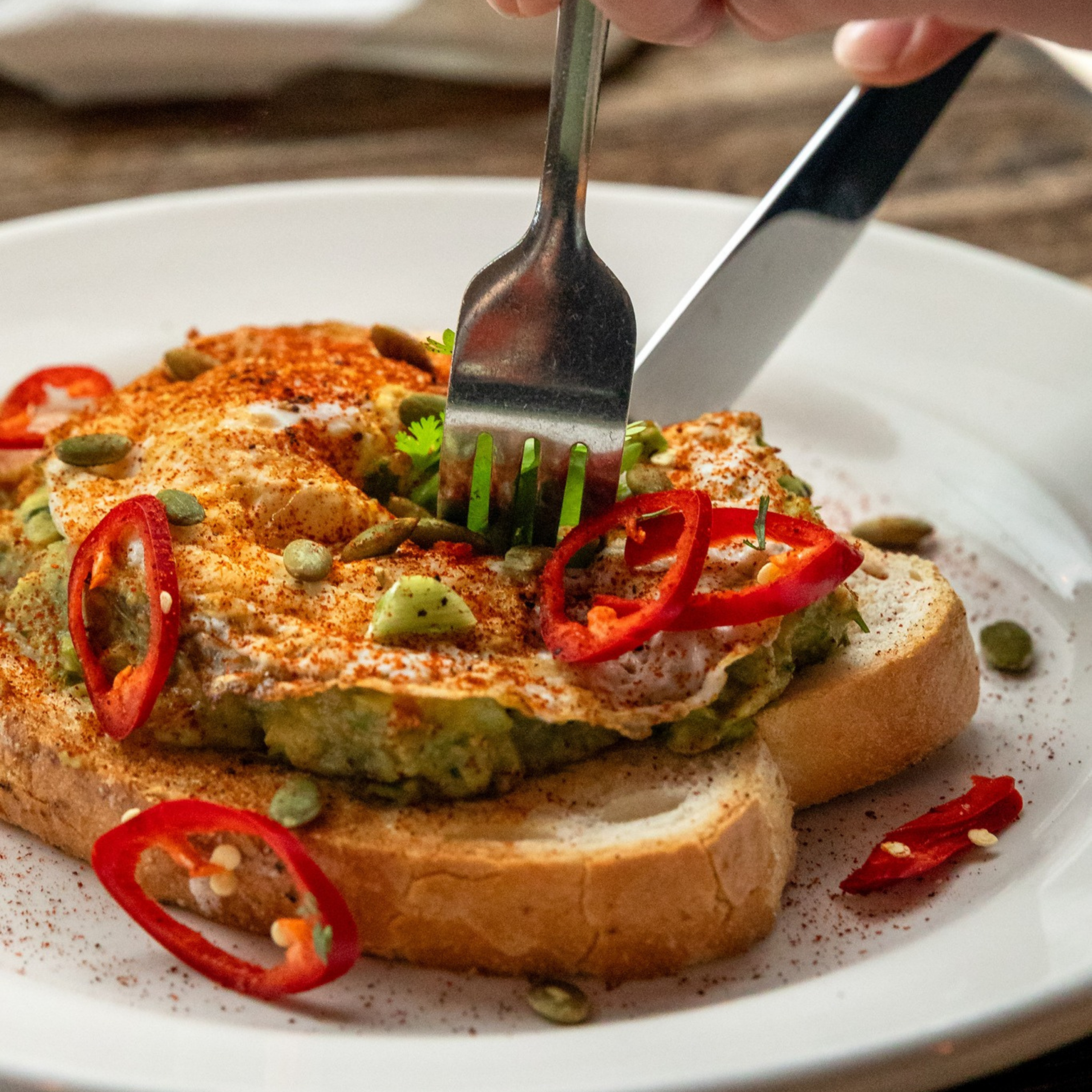 Avocado toast topped with egg, chili slices, and seeds, being cut with fork and knife.