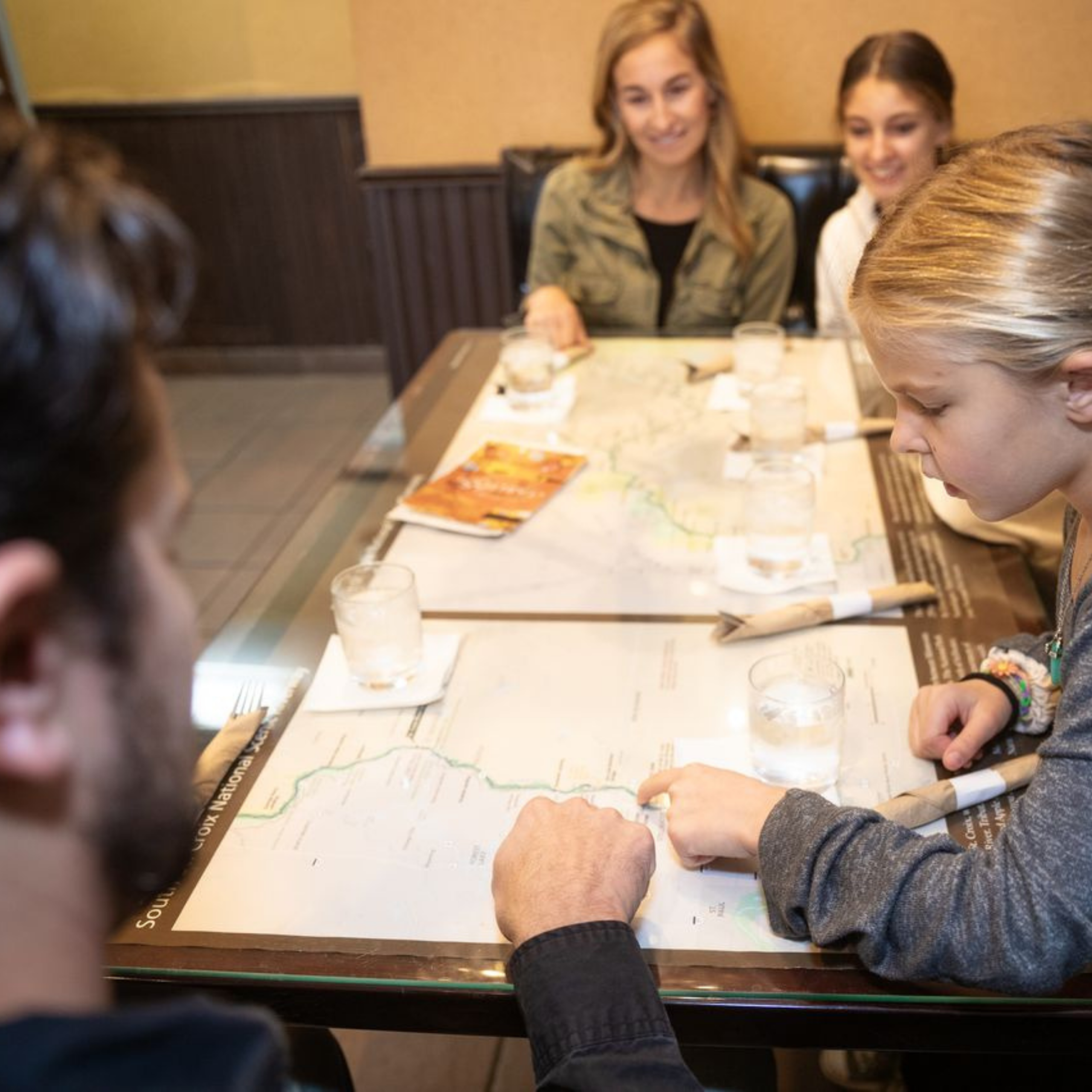 Four people sit at a restaurant table with map placemats, discussing a location.