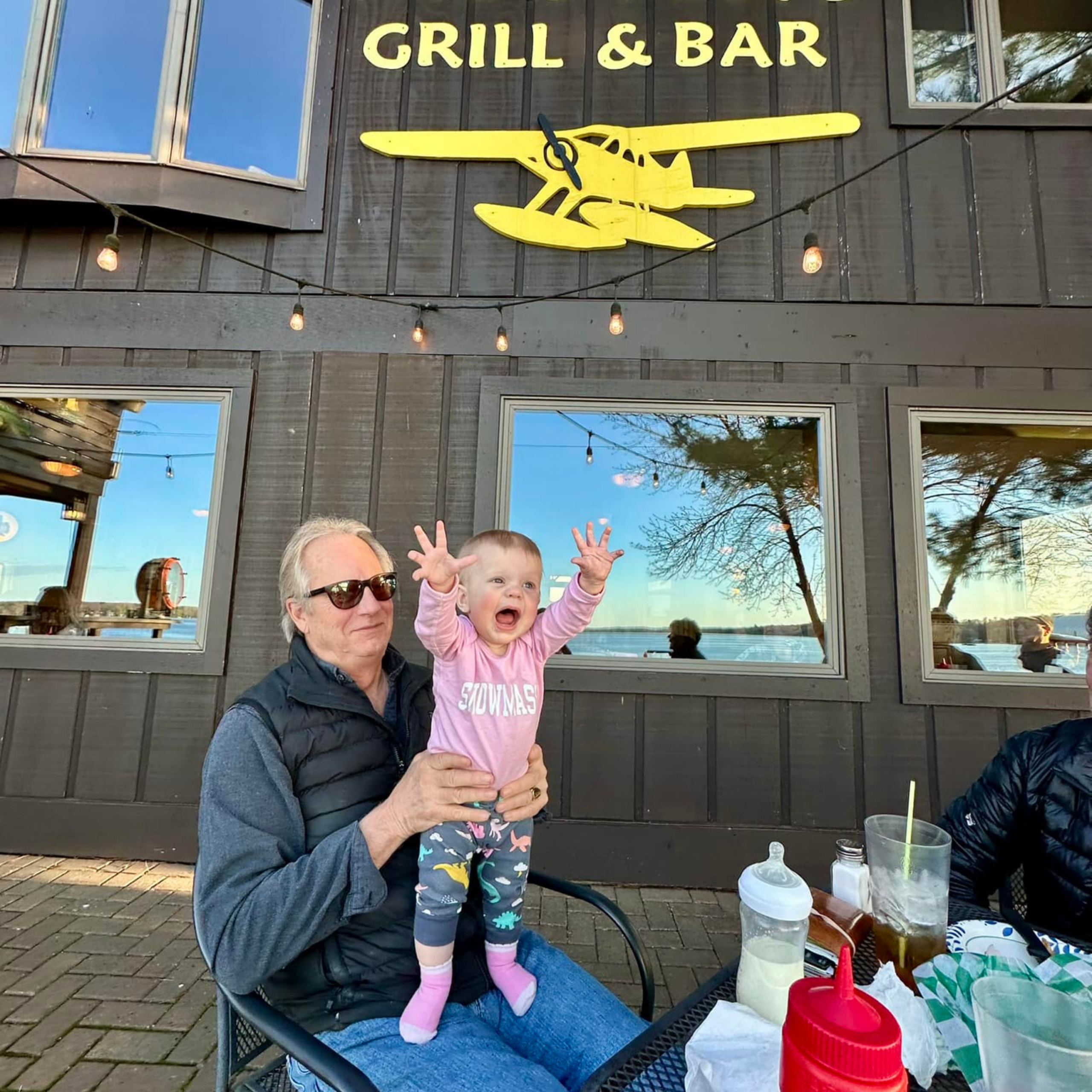 Outdoor dining spaces with a family at Meister’s Grill & Bar. A little girl standing on a man’s lap with an excited expression on her face. 