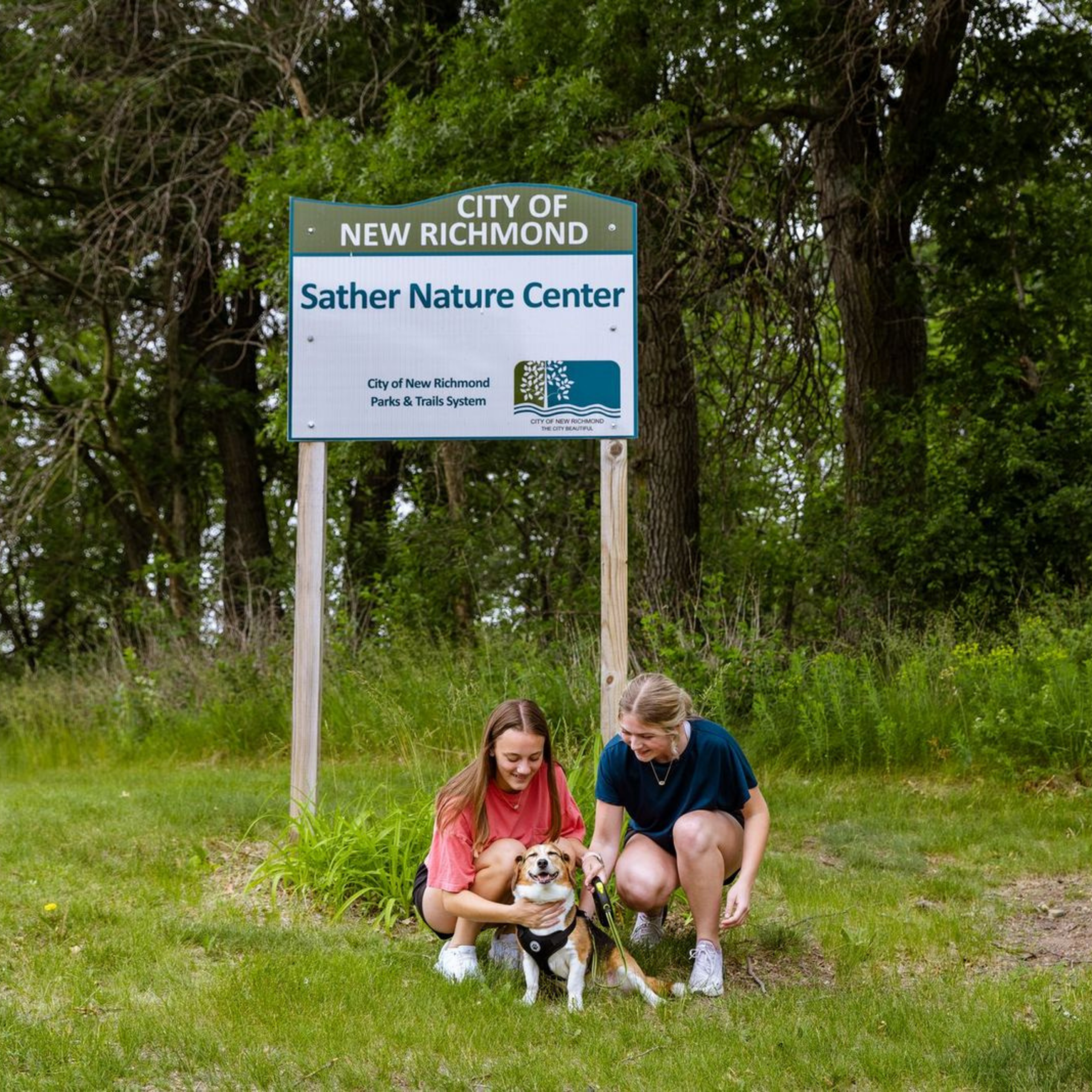 Two women and  a dog sitting in front of Sather Nature Center sign.