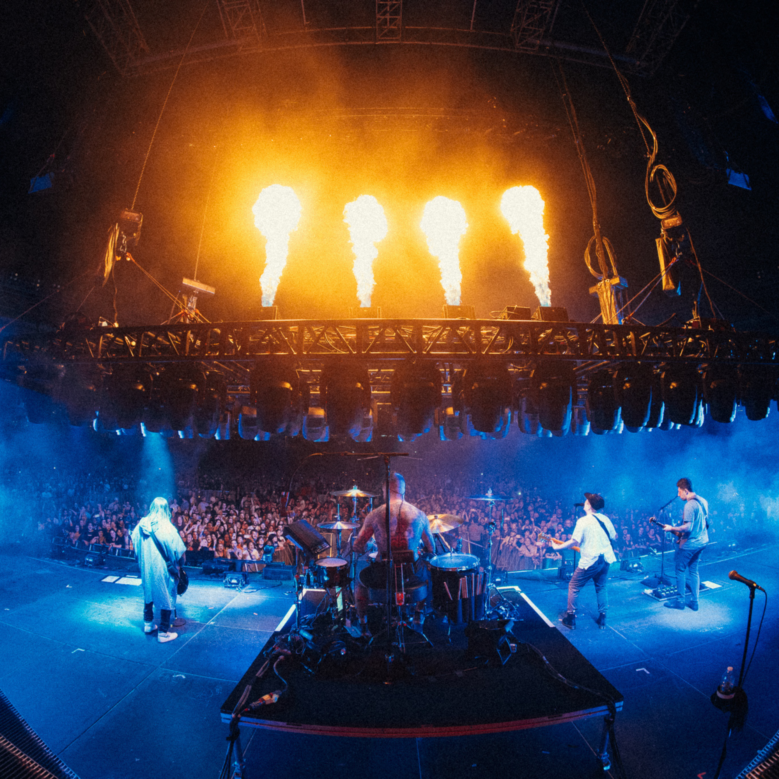 A band is performing for a large crowd. The performance features high-energy production, including blue stage lighting, hazy smoke effects, and pyrotechnic fire blasts in front of the musicians.
