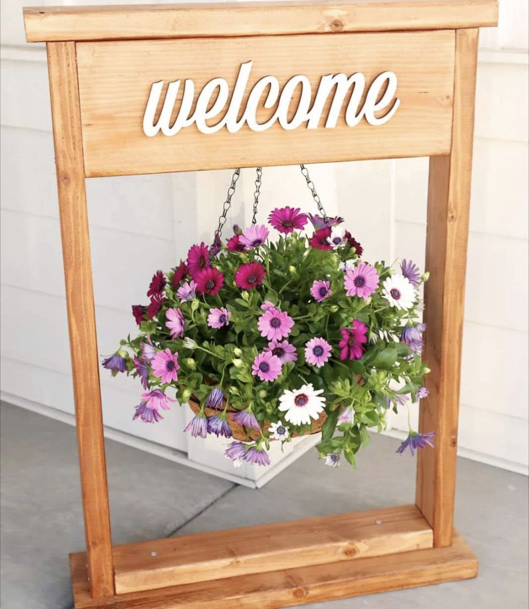 A wooden plant hanger with the words welcome on top in white, with flower hanging from it.
