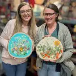 two women holding wood rounds with dried flowers decorating them.