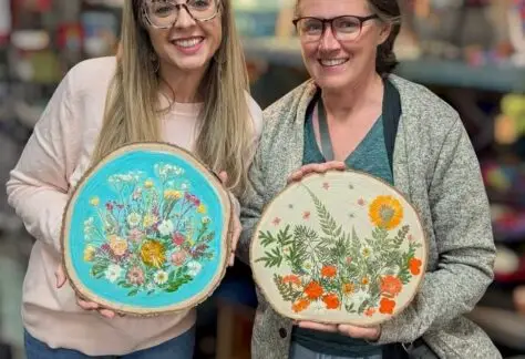 two women holding wood rounds with dried flowers decorating them.