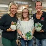 Three women hold painted pumpkins with artificial succulent plants in it