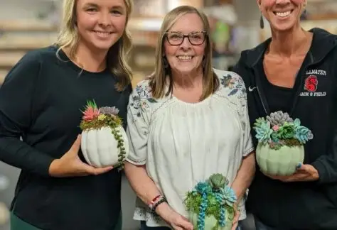 Three women hold painted pumpkins with artificial succulent plants in it