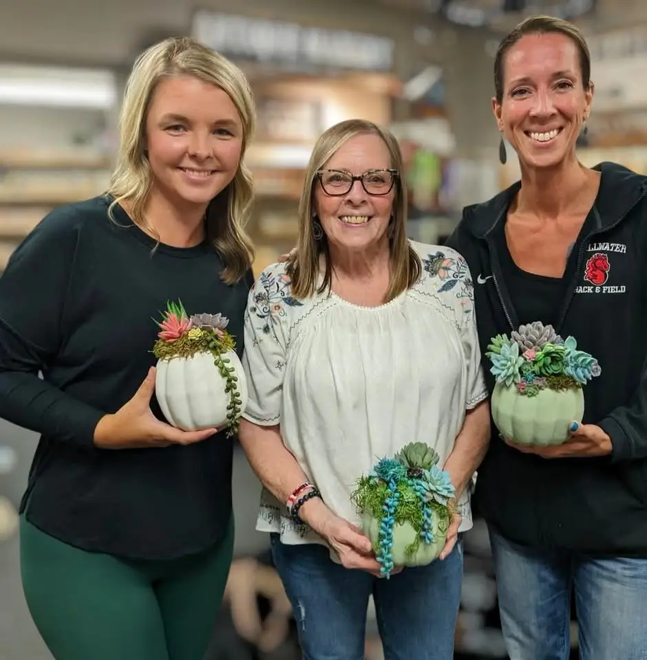 Three women hold painted pumpkins with artificial succulent plants in it